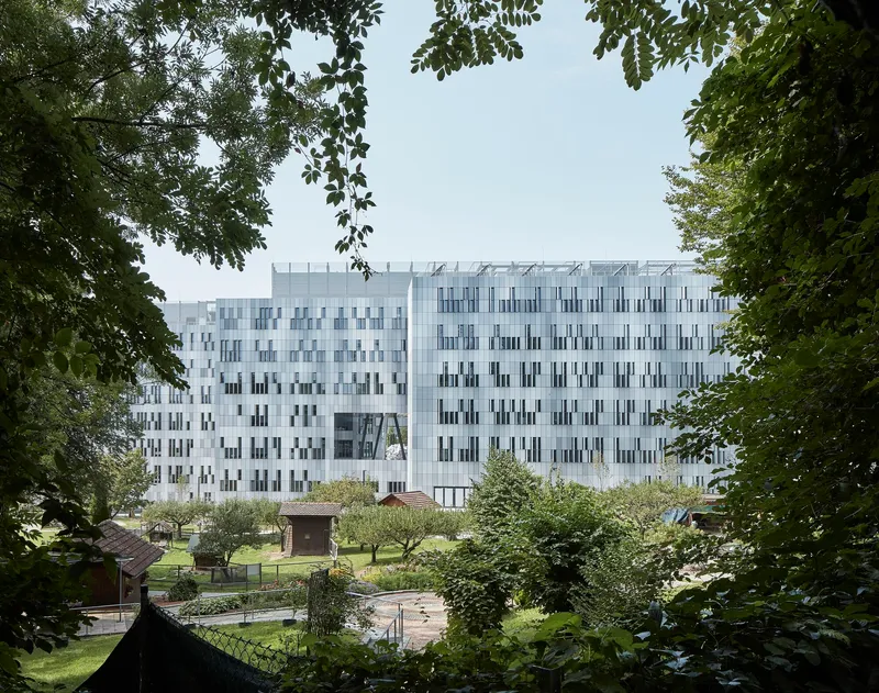White facade with vertical window pattern framed by overhanging tree branches and a garden plaza below
