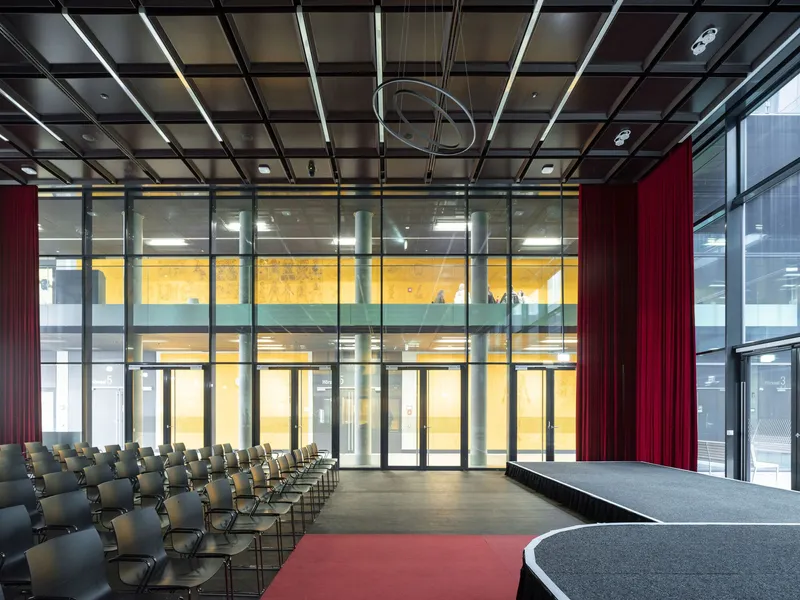 Interior assembly space with red stage curtain and floor-to-ceiling glass overlooking a yellow-lit corridor