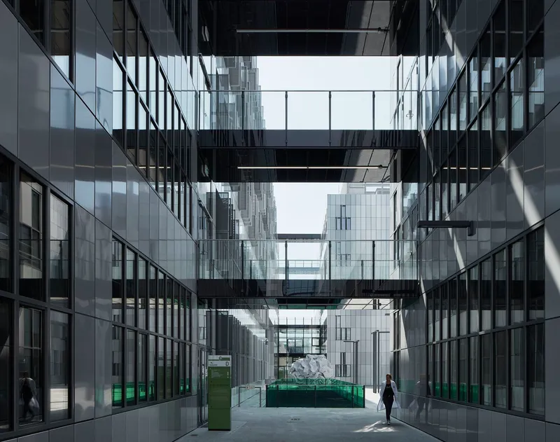 Glazed courtyard with black steel bridges connecting towers as a lone figure walks through sunlit space