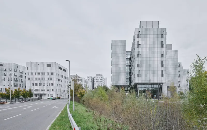 Stepped glass tower beside a roadway with wildflower meadow and residential blocks under overcast sky