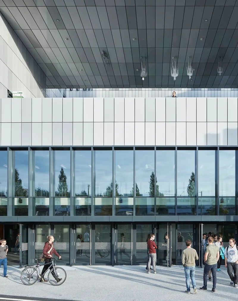 Glazed entrance facade with vertical white panels above and pedestrians gathering on the plaza