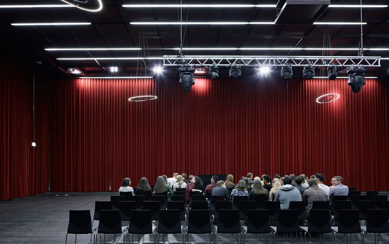 Auditorium with pleated red curtain backdrop and audience seated at rows of black tables