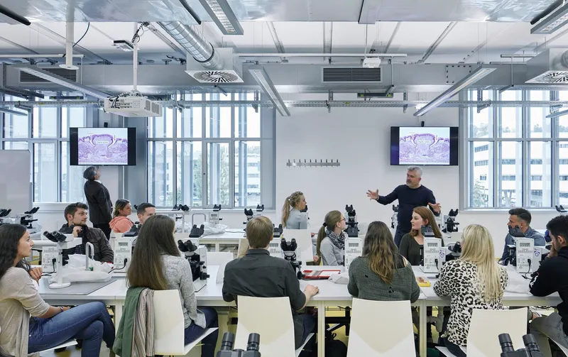 Teaching laboratory with microscopes arranged on white benches beneath exposed ductwork and suspended monitors