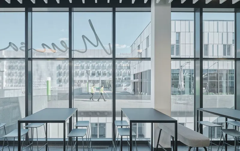 Interior dining area with floor-to-ceiling glazing overlooking a plaza where two people walk past