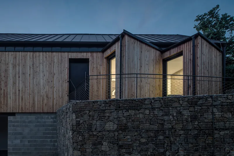 Vertical timber facade with illuminated windows at dusk above gabion retaining wall