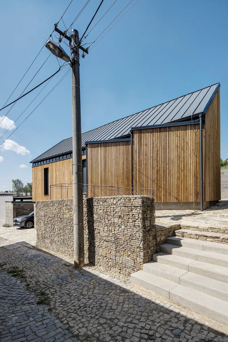Vertical timber facade with gabion stone base and cobblestone paving beneath a clear blue sky