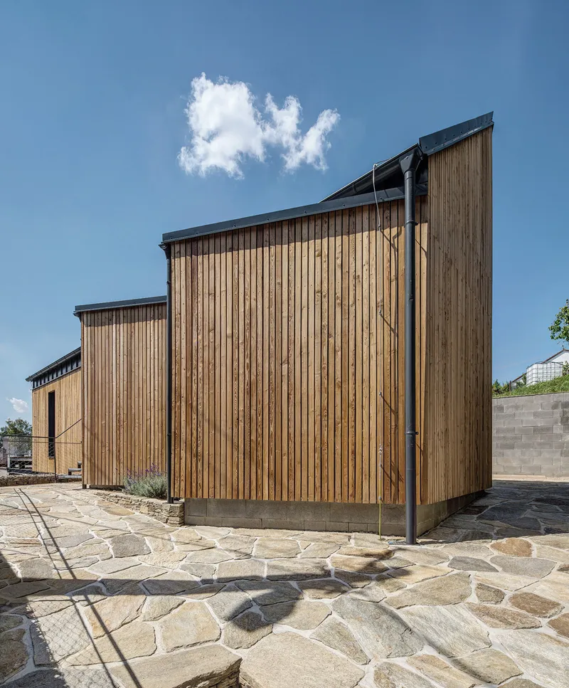 Stepped timber-clad volumes with standing seam metal roofs above irregular stone paving