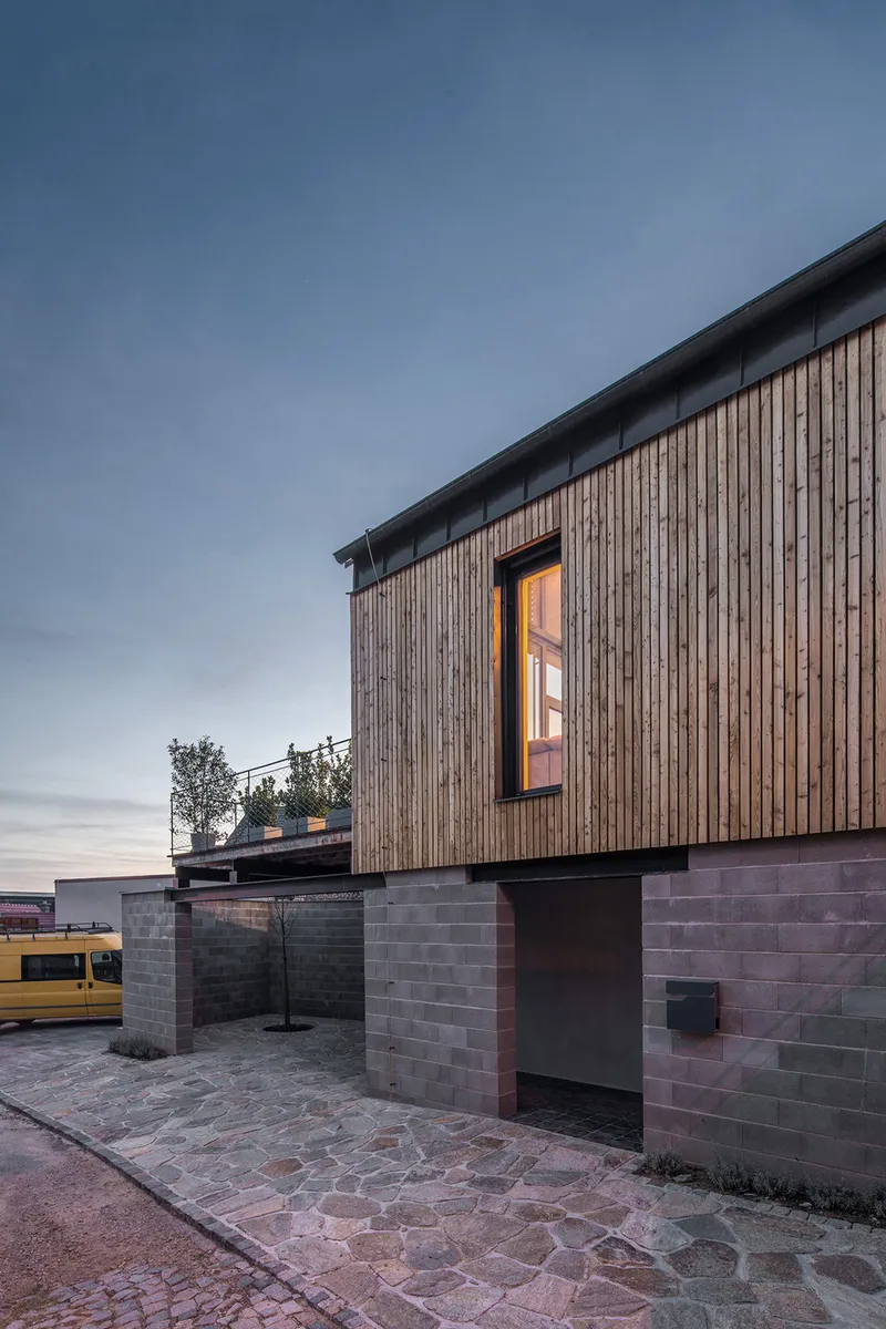 Timber upper volume with illuminated window above concrete block base at dusk