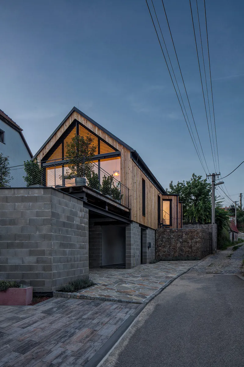 Street view of the timber clad gabled volume above a concrete block base at dusk