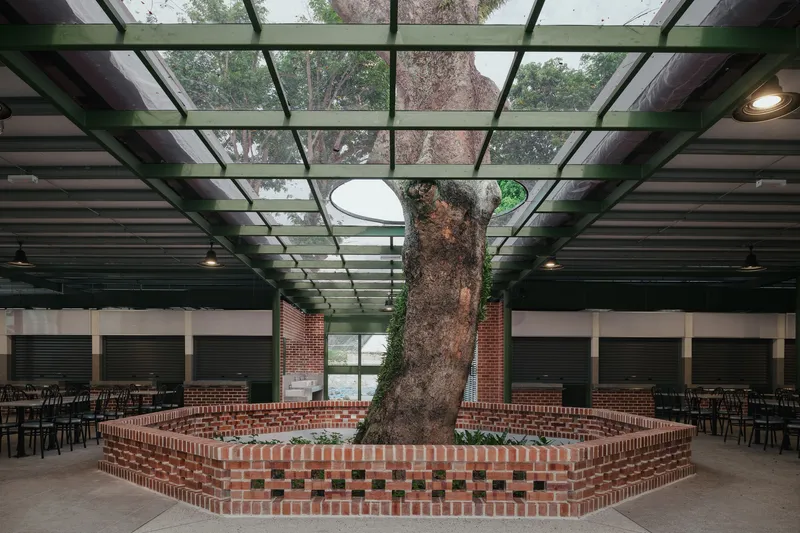 Interior view of a hexagonal brick planter surrounding a preserved tree beneath a glass and steel skylight