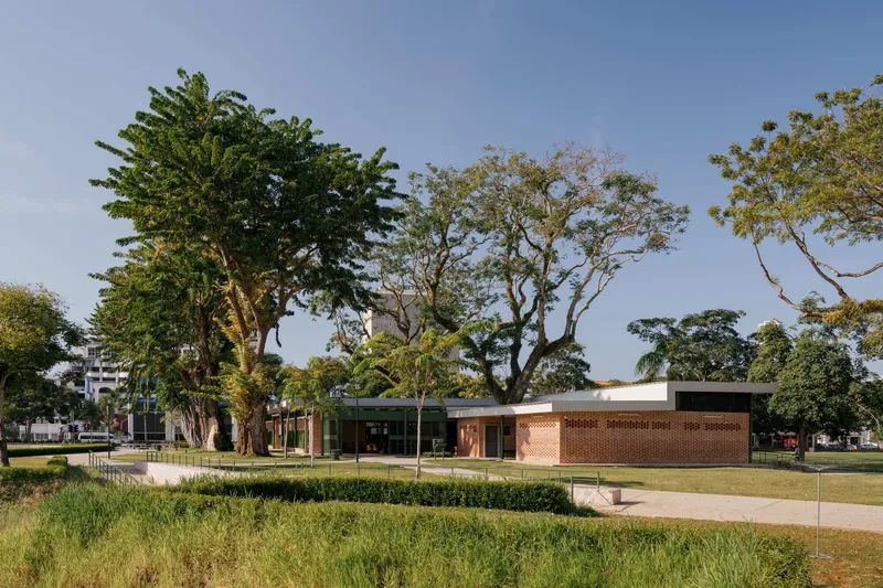 Low-slung brick pavilion with flat roof and clerestory windows set within a landscaped park beneath large trees