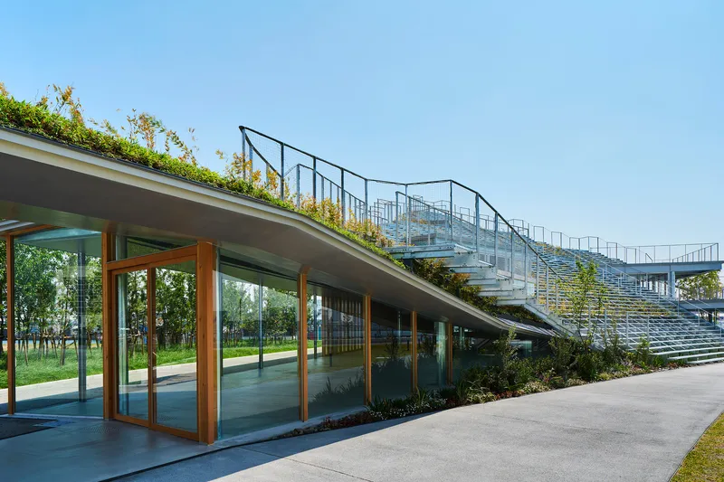 Glass-fronted pavilion beneath planted roof and grandstand seating with metal railings under clear sky