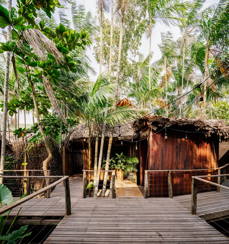 Timber boardwalk approaching thatched-roof pavilion with vertical wood cladding amid coconut palms
