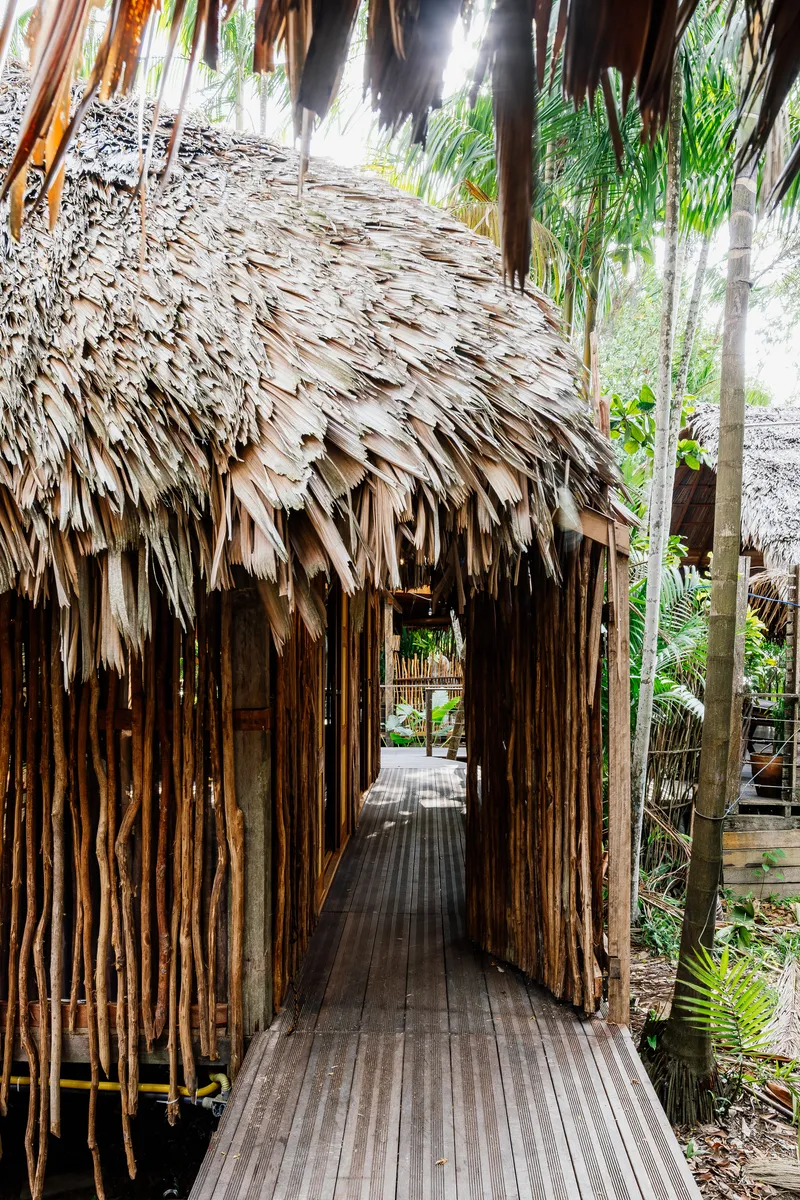 Passageway entrance framed by bundled bamboo walls and layered palm thatch roofing with tropical plantings visible beyond