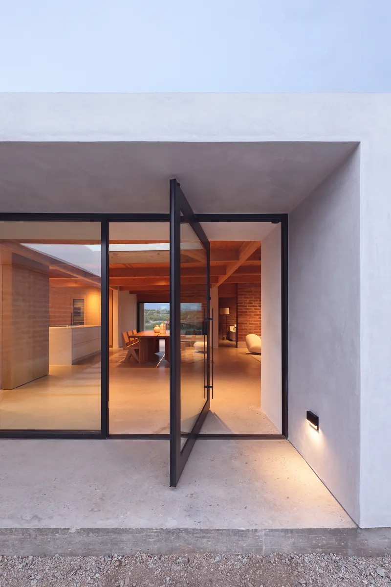 Black steel pivot door swung open beneath white stucco soffit with view through to brick interior