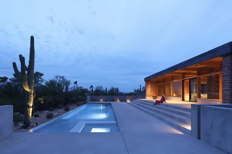 Illuminated pool stretching toward a timber-framed pavilion with saguaro cacti silhouettes at dusk
