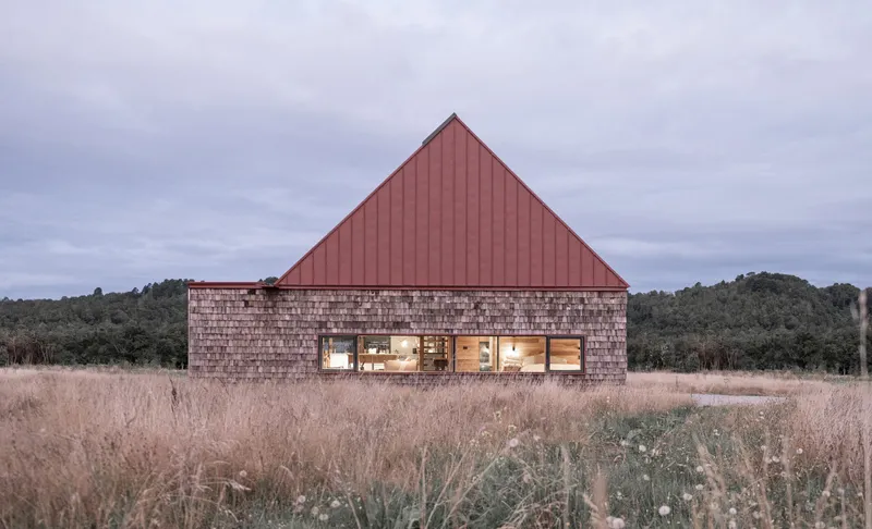 Rear facade with timber shingle walls and red metal gable roof in a dormant grassland at dusk
