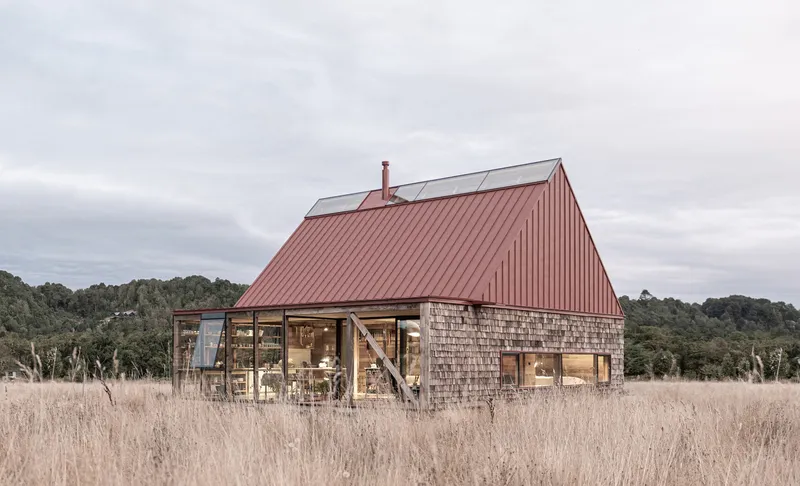 Full view of the shingle and glass house with red metal roof among dry grasses and trees