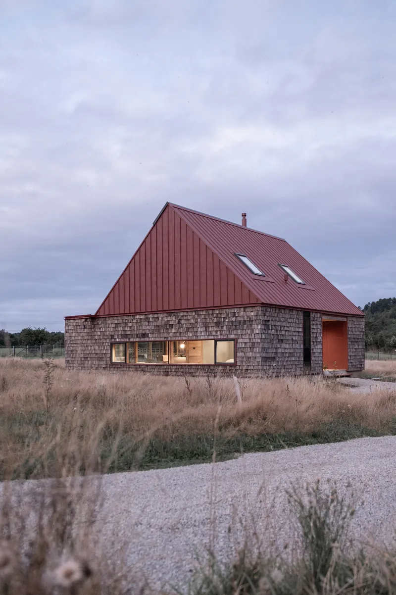 Gabled house with red metal roof and weathered shingle base set in a field at dusk