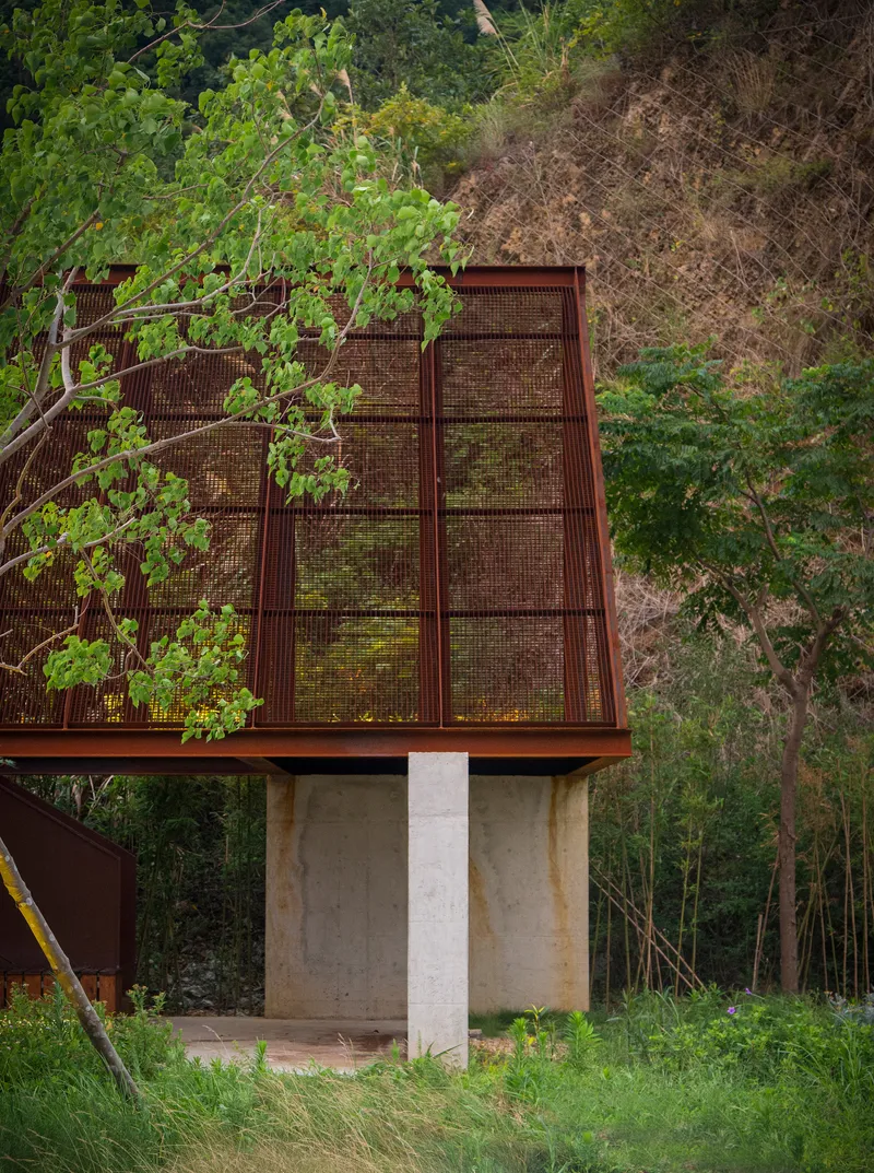 Concrete support beneath a cantilevered mesh-clad roof volume partially screened by young trees