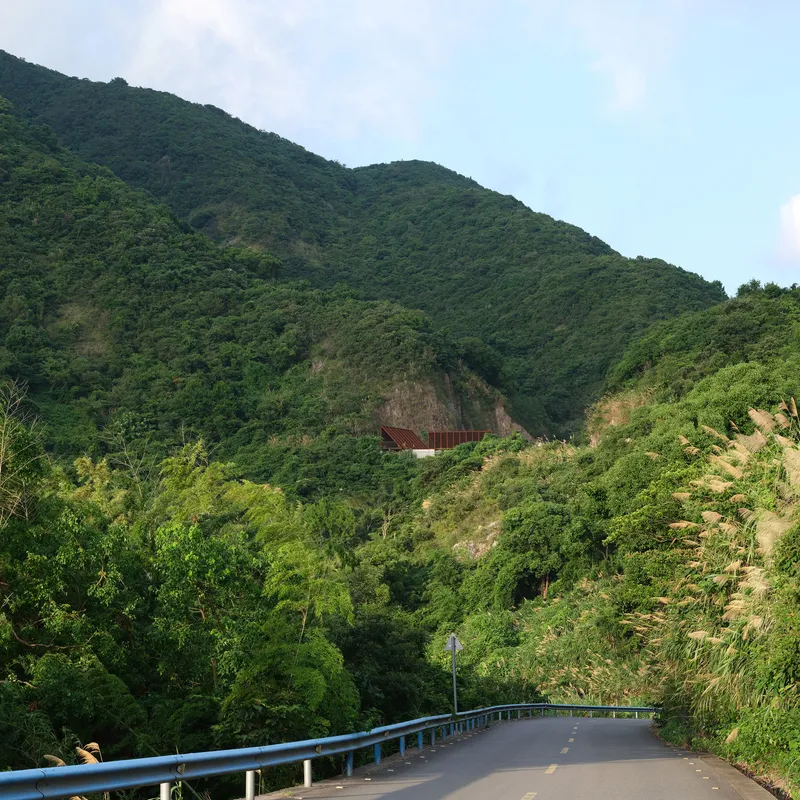Distant view of the structure nestled into the densely forested hillside above a winding road