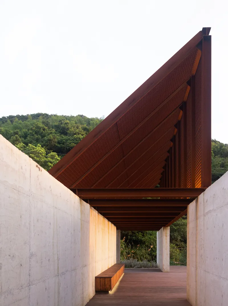 Covered walkway with exposed timber ceiling between concrete walls leading toward the hillside