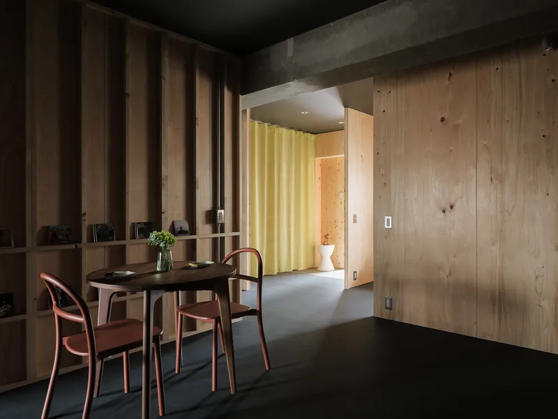 Dining area with plywood shelving unit and yellow curtain visible beyond a concrete beam