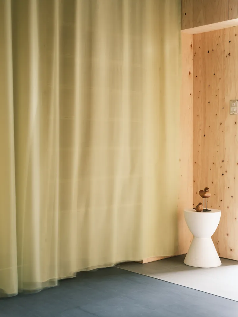 Yellow curtain beside plywood wall with white hourglass stool and small figurine