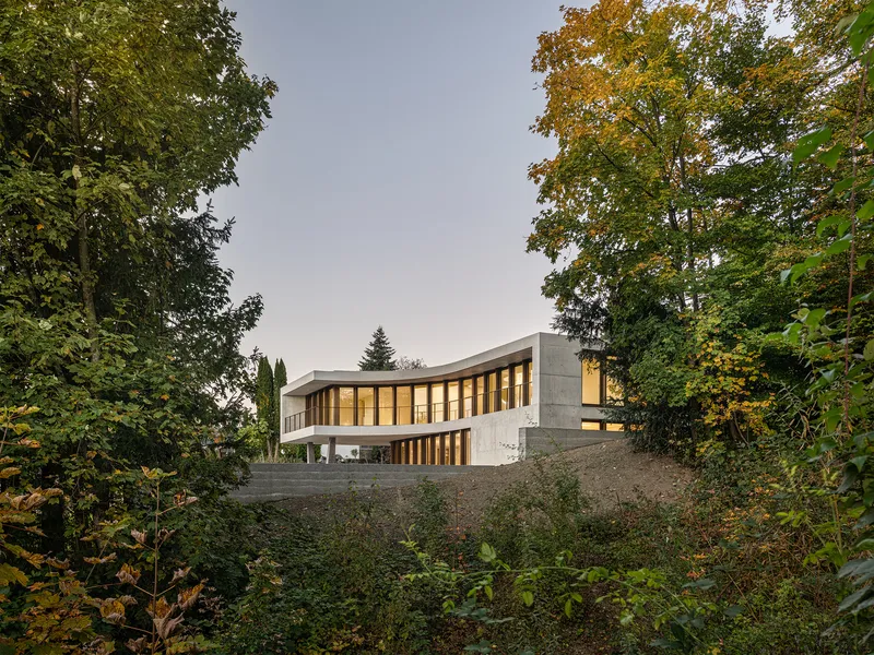 Concrete facade with vertical timber-framed glazing illuminated at dusk from a wooded slope