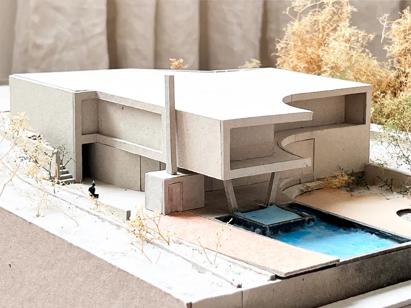 The image showcases an architectural model of a modern house with a minimalist design. The model features a cantilevered structure, a courtyard, and a water fea