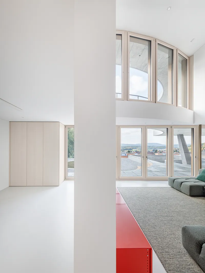 Double-height living space with stacked timber-framed windows and red metal bench overlooking rooftops
