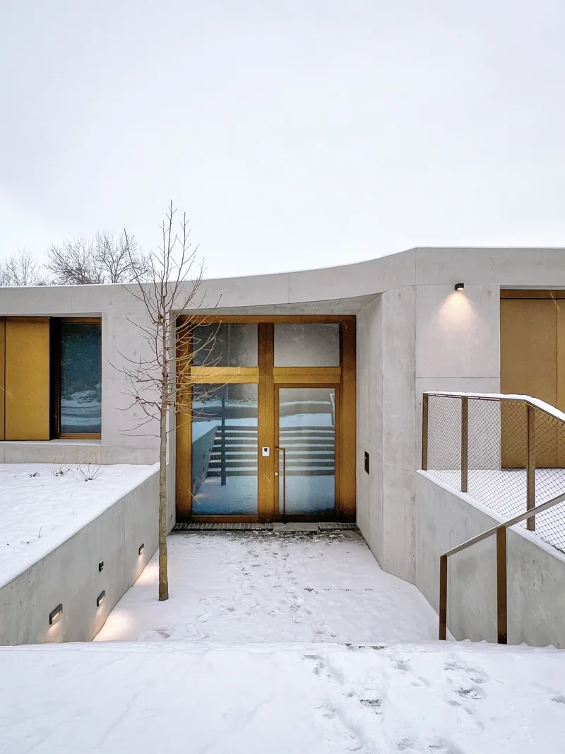 Concrete entrance with timber double doors and metal railing in fresh snow at dusk