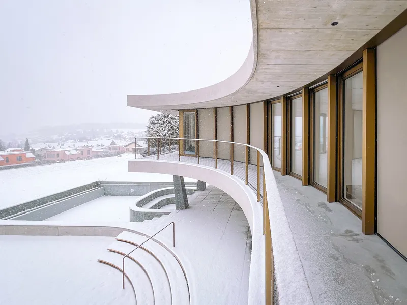 Curved terrace with timber soffit and mesh railing overlooking snow-covered rooftops and hills
