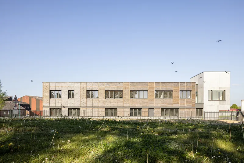 Long light-brick facade with rhythmic window openings above a grassy planted berm with young saplings