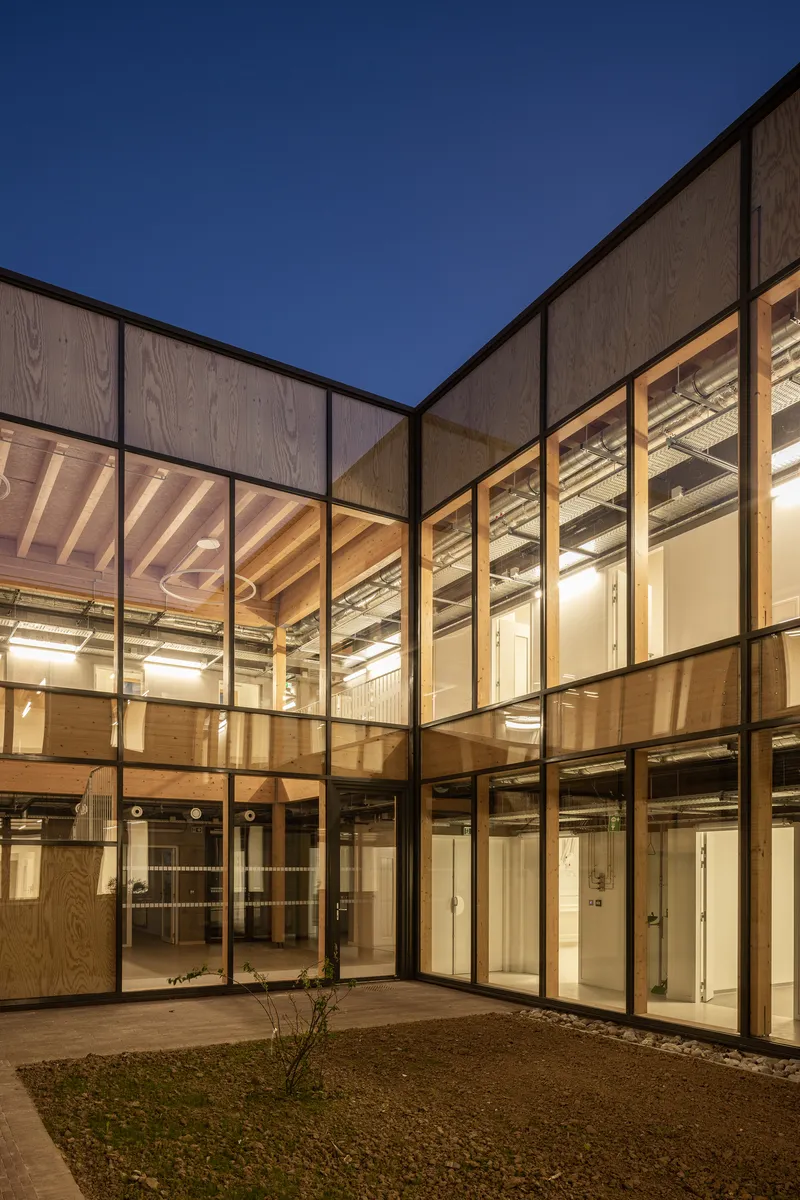 Corner glazed facade revealing timber-framed interior spaces illuminated at dusk under deep blue sky