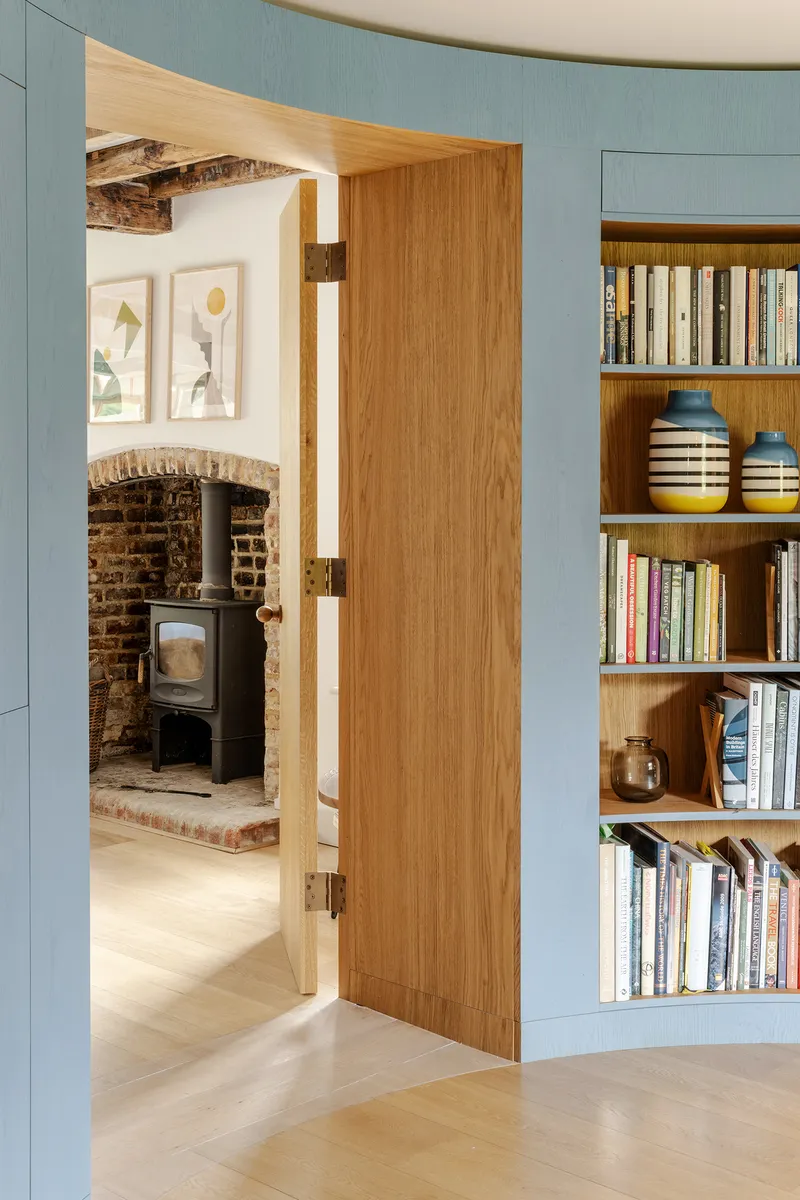 Oak door opening from blue paneled room with built-in shelves to fireplace alcove with exposed brick