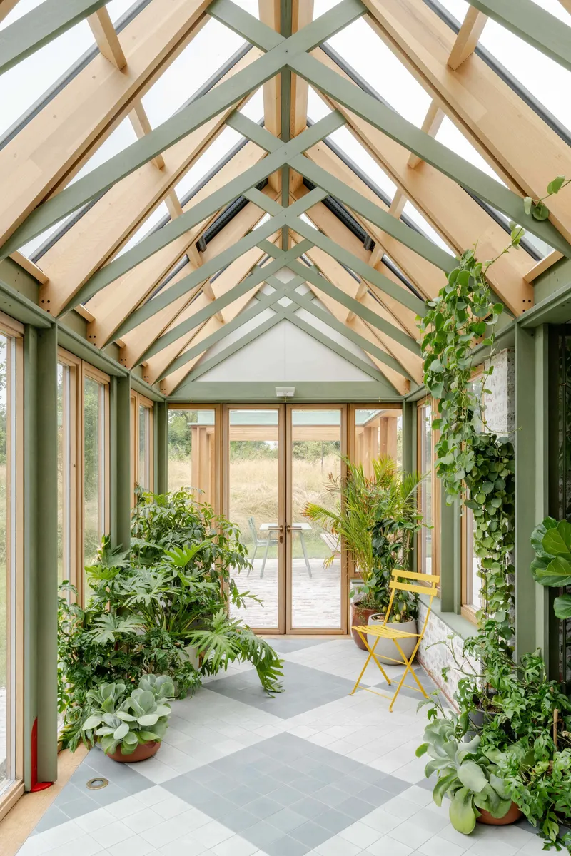 Glazed conservatory interior with exposed timber rafters, skylights and potted plants on grey tile floor
