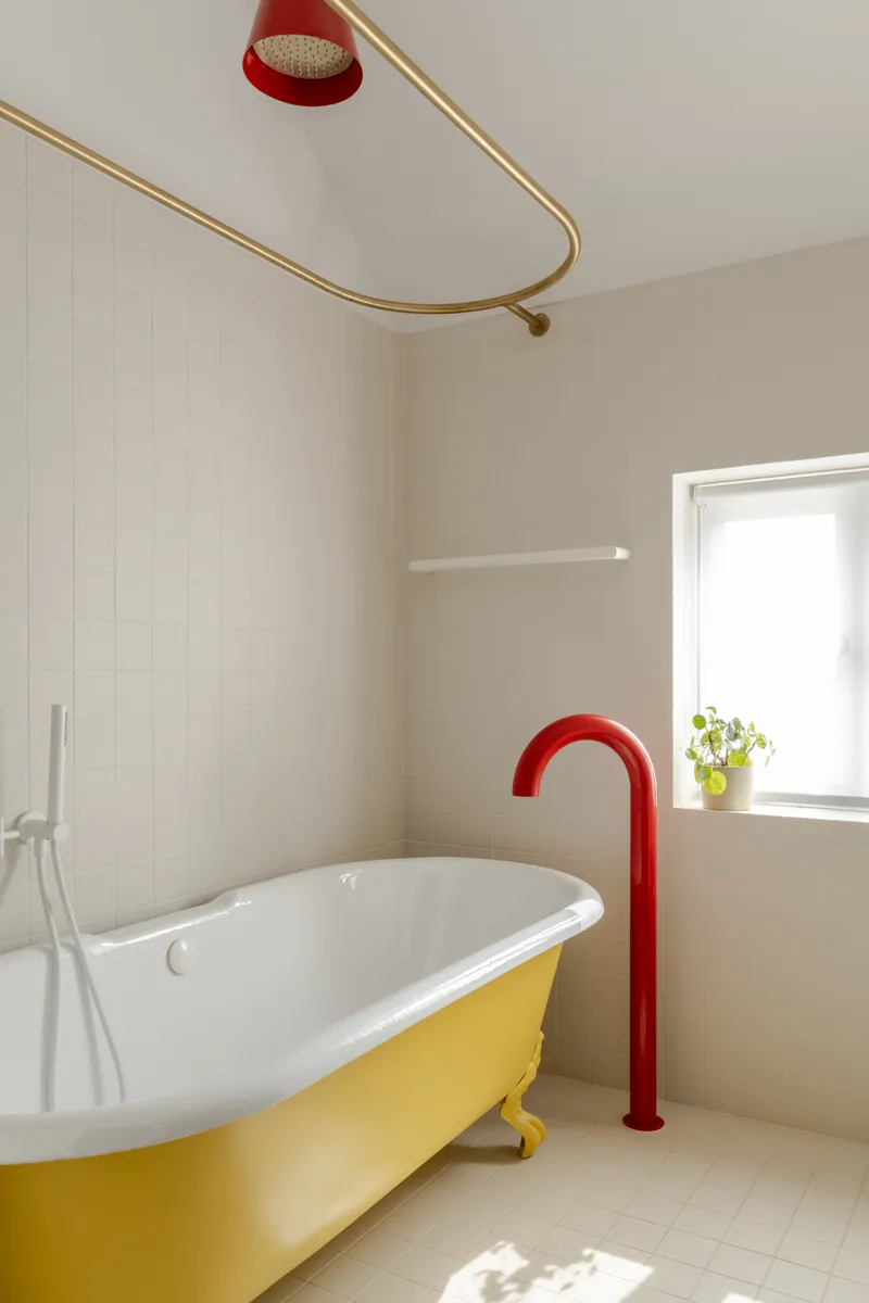 Freestanding tub with yellow base and red floor-mounted tap beneath brass shower rail