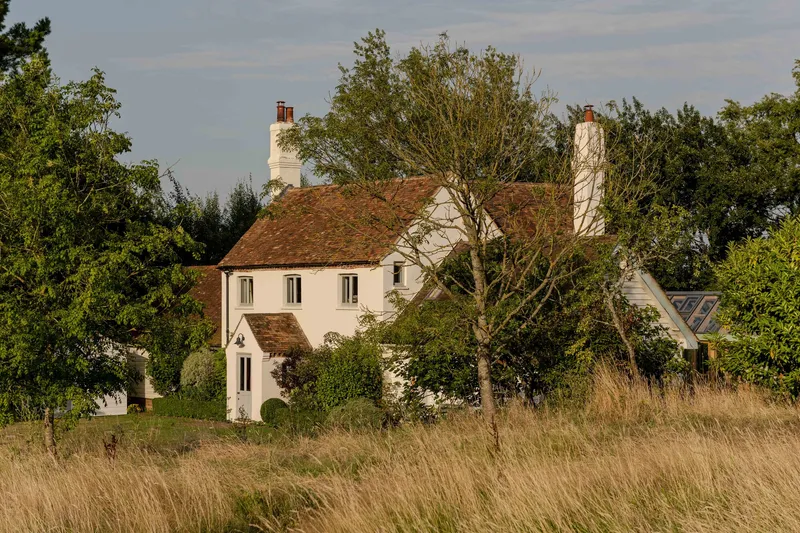 White rendered house with terracotta tile roof amid mature trees and tall grasses at dusk