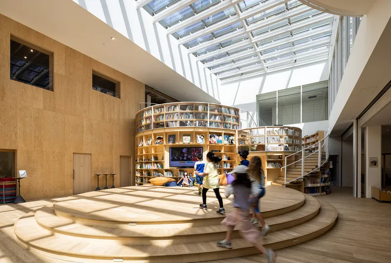 Curved timber platform with circular bookshelf enclosure and children running through sunlit space below skylights