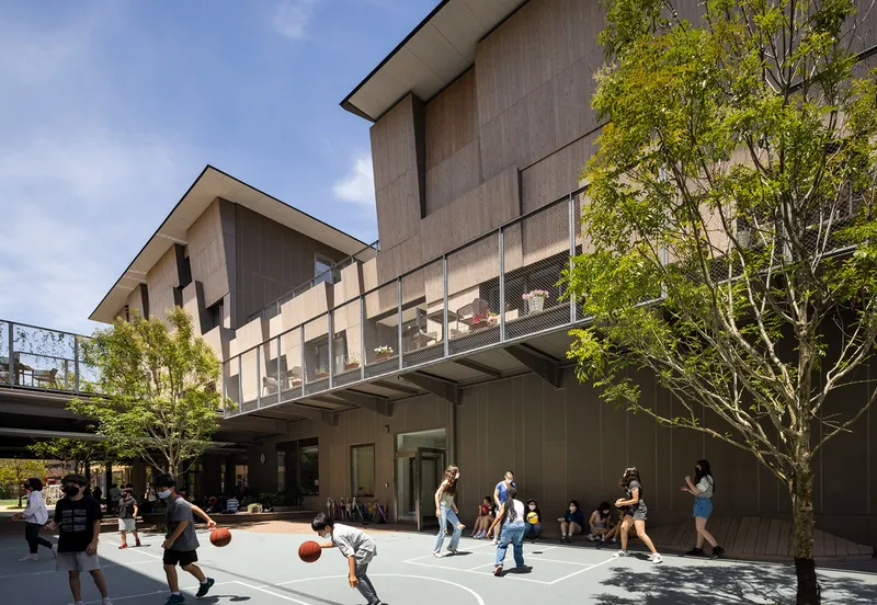 Outdoor basketball court surrounded by timber-clad volumes with an elevated pedestrian bridge and spring foliage