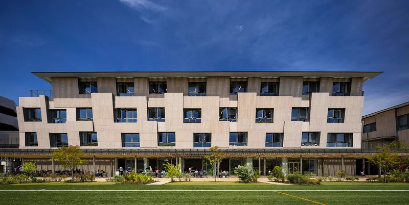 Facade of interlocking pale wood panels with punched windows above a glazed colonnade and lawn