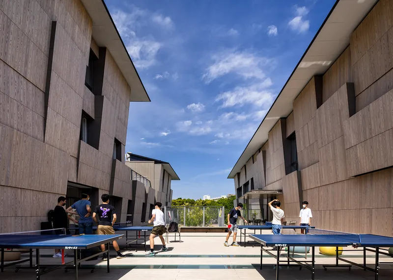 Outdoor courtyard with ping-pong tables between timber-clad buildings under a clear blue sky