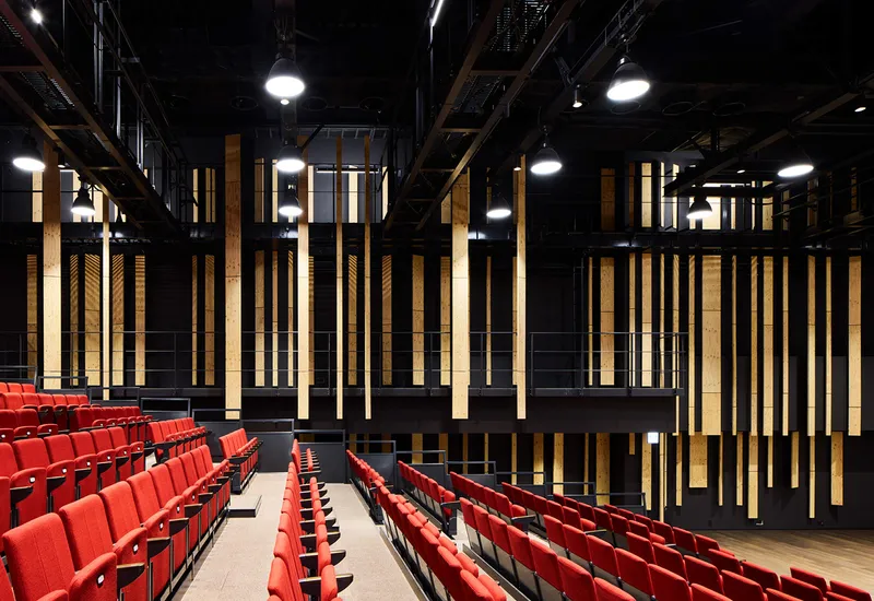 Theater interior with red tiered seating, vertical timber baffles and exposed black technical grid above