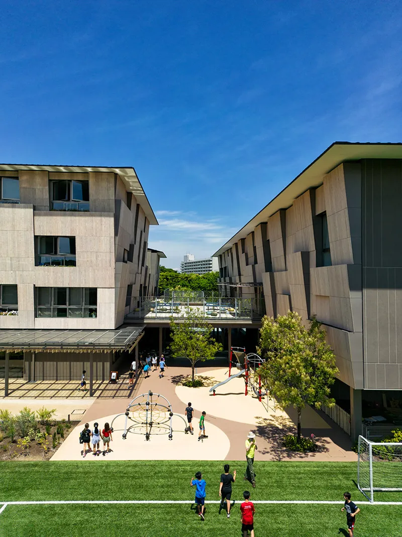 Central courtyard with children playing on turf and pavement between two timber-clad buildings under blue sky