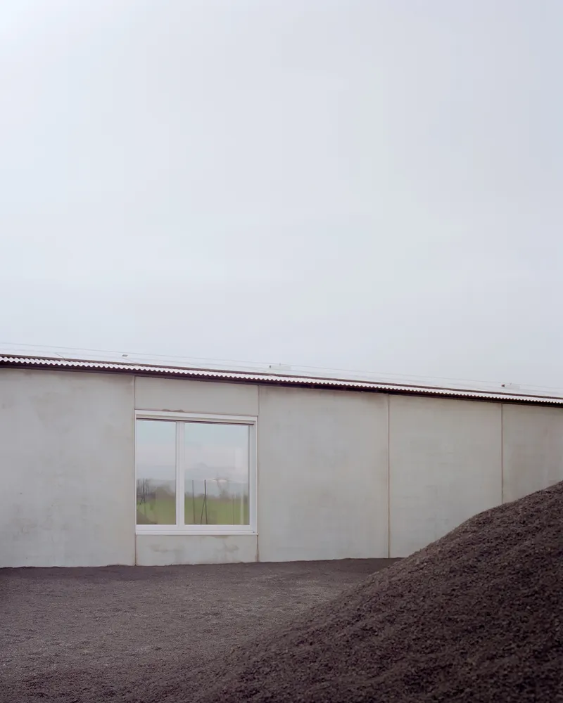 Pale facade with single window and corrugated metal roof beside mound of dark soil