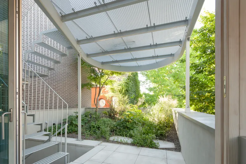 Covered courtyard with translucent corrugated canopy and white metal staircase beside planted bed