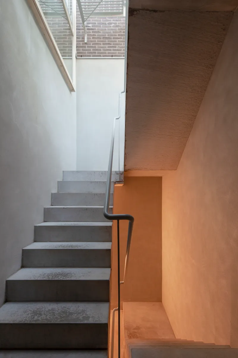 Concrete stair with black metal handrail ascending to glazed opening framing brick wall beyond