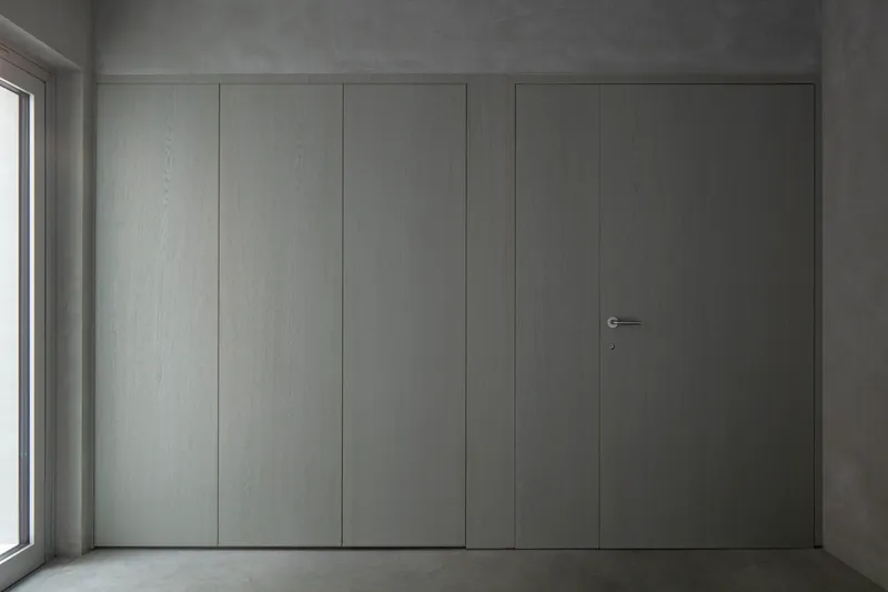 Flush pale grey paneled wall with concealed door and minimal hardware beside a window