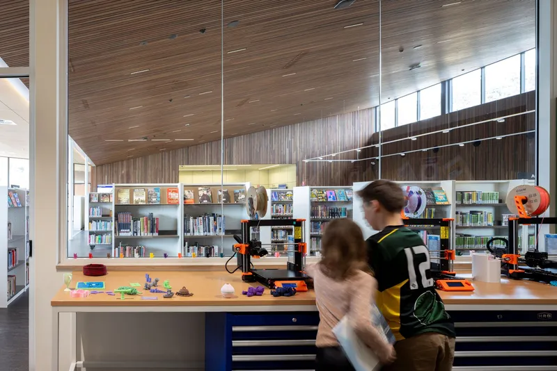 Makerspace workbench with 3D printers and blurred figures moving through the timber-clad room